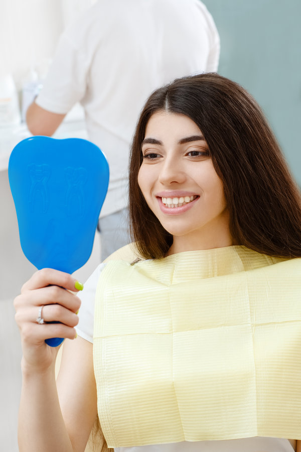 Teeth problems are in the past. Young attractive woman checking her smile looking in the mirror sitting in a dental chair professional dentist on the background