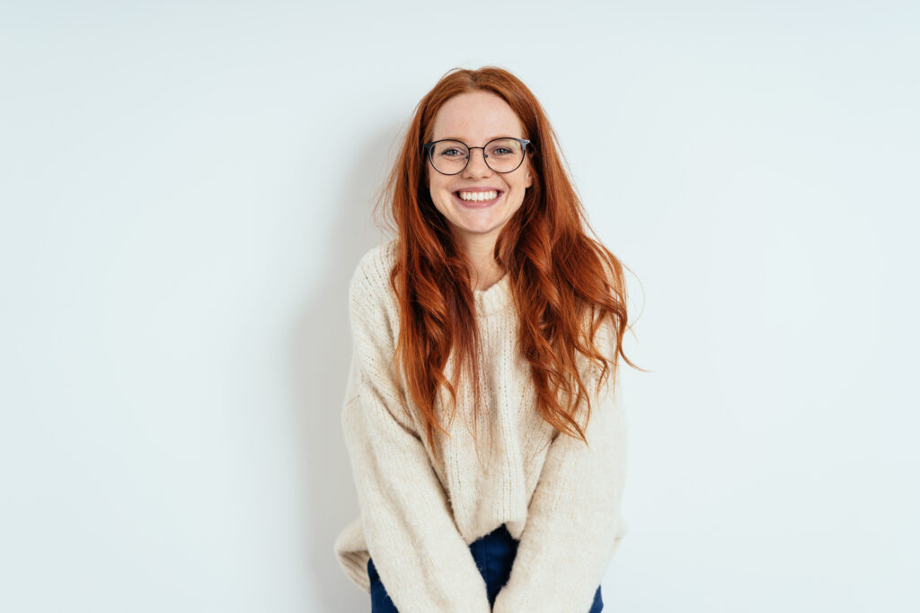 Smiling friendly young woman with long red hair wearing spectacles looking at the camera with a vivacious smile against a white interior wall with copy space