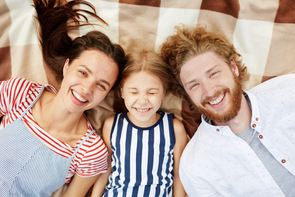 Carefree young man, woman and little girl looking at camera while relaxing on plaid