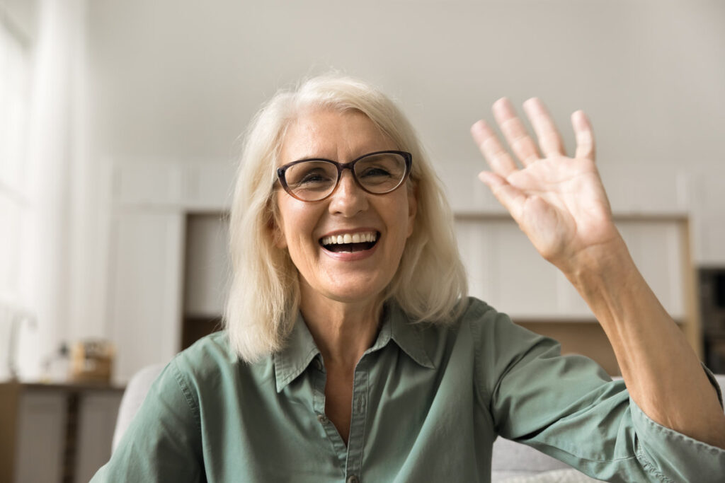 Cheerful beautiful blonde elder woman in stylish eyeglasses talking on online conference call, waving hi hand, saying hello, laughing, looking at camera with toothy smile. Head shot portrait