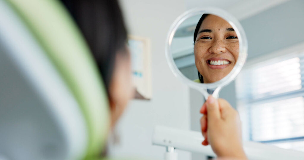 Dental, smile and happy woman with mirror check at a dentist for teeth whitening results. Mouth, oral care and Asian female client with tooth, cleaning or routine checkup in Tokyo with satisfaction.