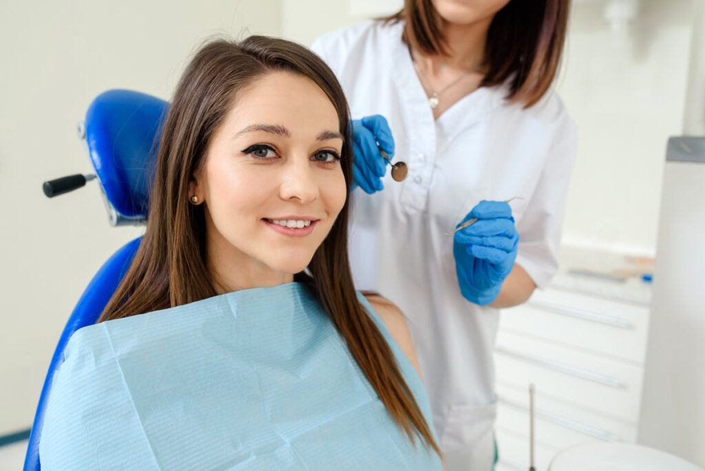 Young happy smiling woman patient in a dental clinic is receiving a dental treatment and looking at the camera. The dentist with dental equipment are behind